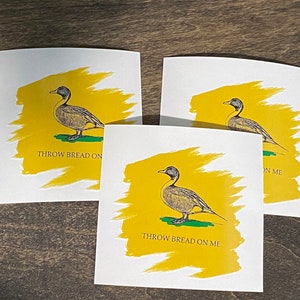 Puede incluir: Tres pegatinas cuadradas blancas con fondo amarillo y una ilustración en blanco y negro de un pato. El texto "Throw Bread On Me" está impreso en cada pegatina.