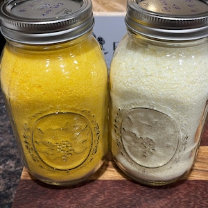 May include: Two glass mason jars with silver lids, filled with yellow and white powders. The jars have embossed designs and are on a wooden surface. The jar on the left is labeled "Egg Yolk".