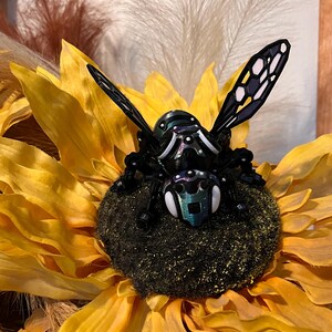 May include: A black and iridescent bee with white accents sits on a yellow sunflower. The bee has detailed wings and a textured body.