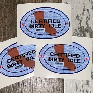 Könnte beinhalten: Drei weiße Aufkleber mit einem blauen ovalen Rand und einer braunen Umrandung des Bundesstaates Kalifornien. Der Oval enthält den Text "CERTIFIED DIRTY IDLE" in schwarzer Schrift mit einem roten Stern auf jeder Seite des Textes. Der Text "DIRTY IDLE" wird in kleinerer Schriftgröße um den ovalen Rand wiederholt.