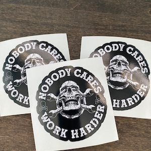 以下が含まれることがあります： 頭蓋骨と交差したレンチが描かれた白黒のステッカーが3枚。ステッカーには「Nobody Cares Work Harder」という文字が書かれています。