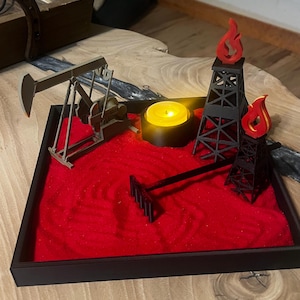 May include: A miniature oil rig diorama with a red sand base, a small candle, and a rake. The scene includes a model oil pump jack and two black oil derricks with red flame accents, all set within a black tray.