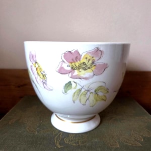 May include: A white porcelain bowl featuring a delicate watercolour-style illustration of pink and yellow wild roses with green leaves. The bowl has a rounded shape, a pedestal base, and a thin gold band accenting the rim of the base.