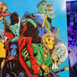 May include: A wood-burned illustration of a group of cartoon characters. The characters are a green monster with a large mouth, a woman with black hair and green skin, a yellow robot with a helmet, and a blue alien with a helmet. The background is blue.