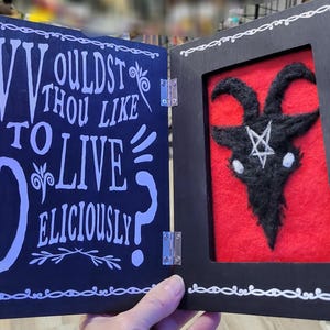 May include: A dark wooden book-shaped box with the text "WOULDST THOU LIKE TO LIVE DELICIOUSLY?" on the left side. The right side features a black goat head with a white pentagram on a red background. The box is hinged.