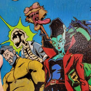 May include: A hand-painted wooden panel with a comic book style illustration of four characters. The characters are a green-skinned woman with red hair, a skeleton, a man with a beard, and a creature with a long tongue. The background is blue with a green and yellow starburst.