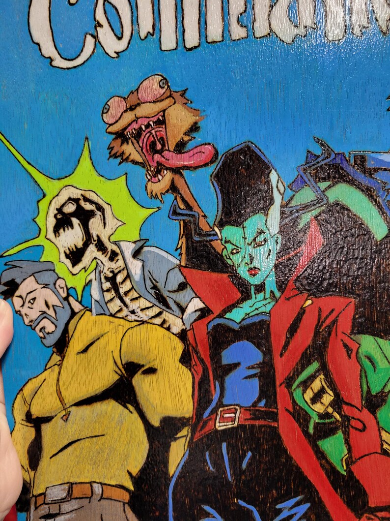 May include: A hand-painted wooden panel with a comic book style illustration of four characters. The characters are a green-skinned woman with red hair, a skeleton, a man with a beard, and a creature with a long tongue. The background is blue with a green and yellow starburst.