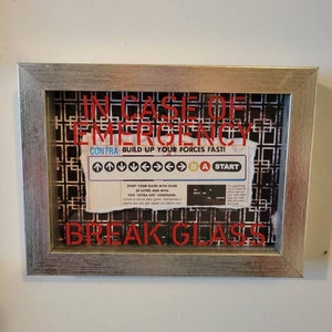May include: A framed image with a red background and the text "In Case of Emergency" in white letters. The image also includes a black and white image of a video game screen with the text "CONTRA BUILD UP YOUR FORCES FAST!" and the words "BREAK GLASS" in red letters.