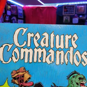 May include: A blue wooden sign with the words "Creature Commandos" in white lettering. The sign features a cartoon illustration of a green monster with a long tongue and a brown creature with a long neck and a long tongue.