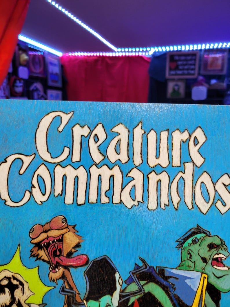 May include: A blue wooden sign with the words "Creature Commandos" in white lettering. The sign features a cartoon illustration of a green monster with a long tongue and a brown creature with a long neck and a long tongue.