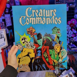 May include: A blue wooden sign with the words "Creature Commandos" in white lettering. The sign features a comic book style illustration of five characters, including a zombie, a vampire, a werewolf, a monster, and a human.