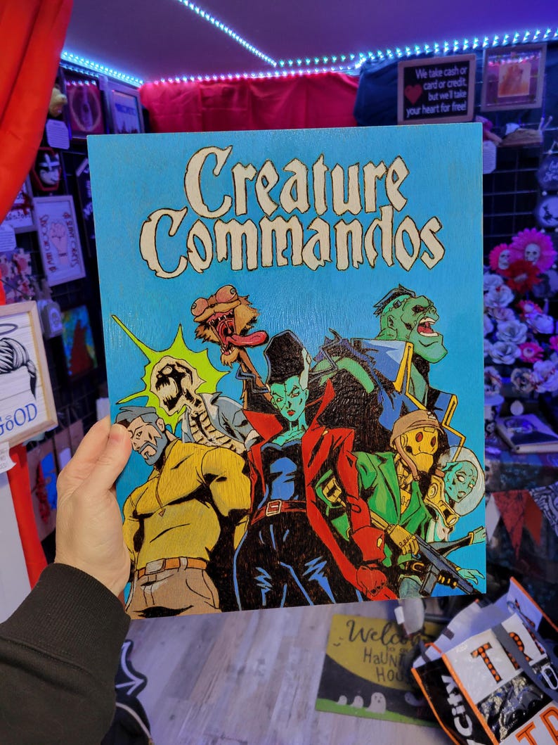 May include: A blue wooden sign with the words "Creature Commandos" in white lettering. The sign features a comic book style illustration of five characters, including a zombie, a vampire, a werewolf, a monster, and a human.