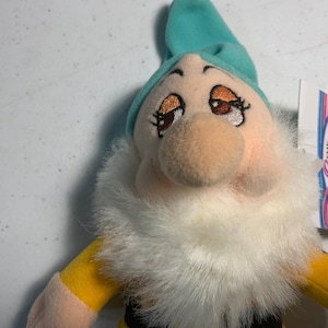 May include: A plush doll of a dwarf character, featuring a light blue hat, a large nose, and a white beard. The dwarf has rosy cheeks, brown eyes, and yellow sleeves. The doll is a collectible item.