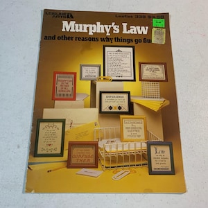 May include: A vintage leaflet titled "Murphy's Law" with various framed cross-stitch samplers. The samplers feature humorous sayings and quotes, including "Assumption is the mother of all screw ups" and "Experience is something you don't get until just after you need it."