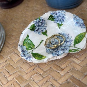 May include: A small, white, decorative dish with a floral hydrangea design in blue and green. Two gold hoop earrings rest inside the dish. The dish sits on a woven, light brown surface.