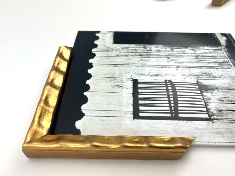 May include: A gold-colored, textured wood frame with a black and white photograph of a wooden building with a window and a gate. The frame has a scalloped edge.