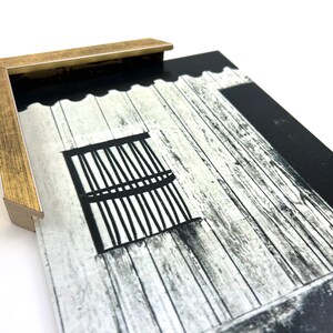 Puede incluir: Una fotograf&iacute;a en blanco y negro de un edificio de madera con una ventana. La ventana tiene barrotes negros y est&aacute; enmarcada en blanco. El edificio est&aacute; enmarcado en un marco dorado.