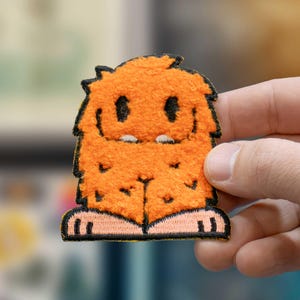 May include: An orange, fuzzy monster patch with black eyes, a small smile, and two white teeth. The patch has black trim and tan feet. The patch is held by a hand.