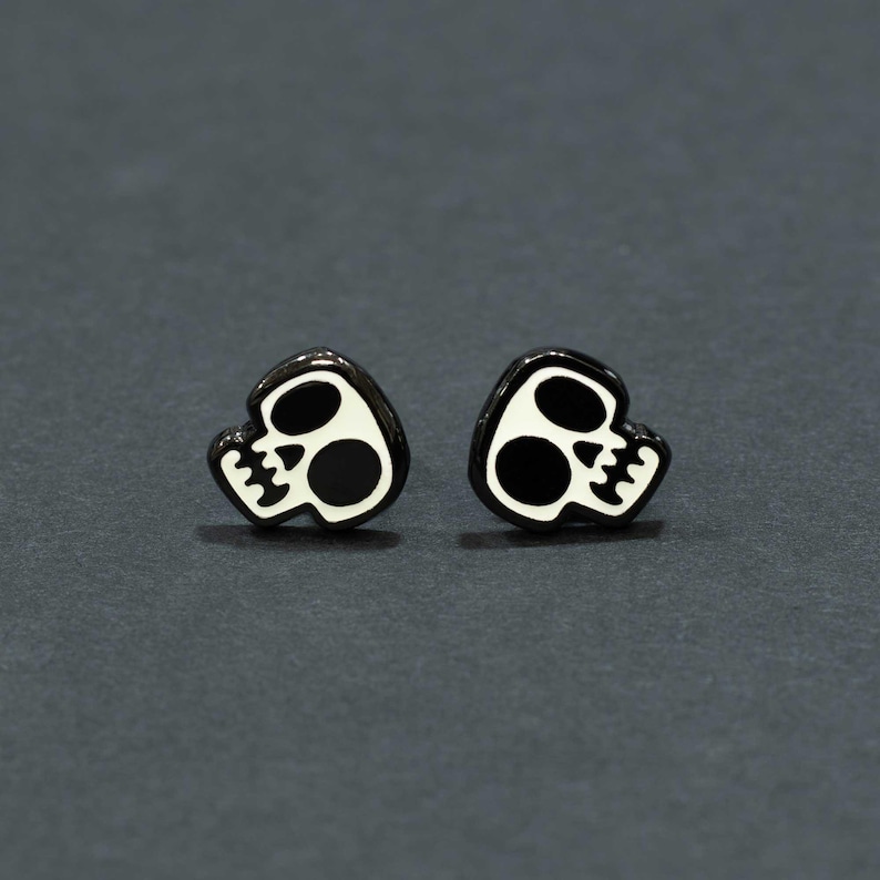 May include: A pair of black and white skull-shaped stud earrings. The earrings feature a glossy black outline with a white skull design, including eye sockets and a jawline. The earrings are set against a dark gray background.