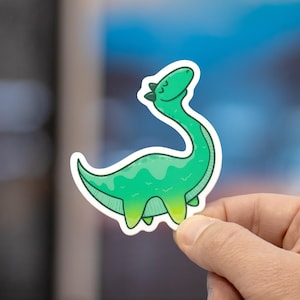 May include: A die-cut sticker featuring a cartoon long-necked dinosaur in shades of bright green and lime green. The dinosaur has a smooth, rounded body, a curved neck, and a small head with closed eyes, outlined in a thick white border.