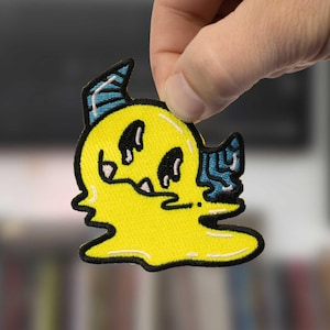 Puede incluir: Parche bordado amarillo con un personaje de dibujos animados con forma de fantasma con ojos y boca negros. El personaje tiene detalles en azul y un borde negro. El parche está sujeto por una mano.