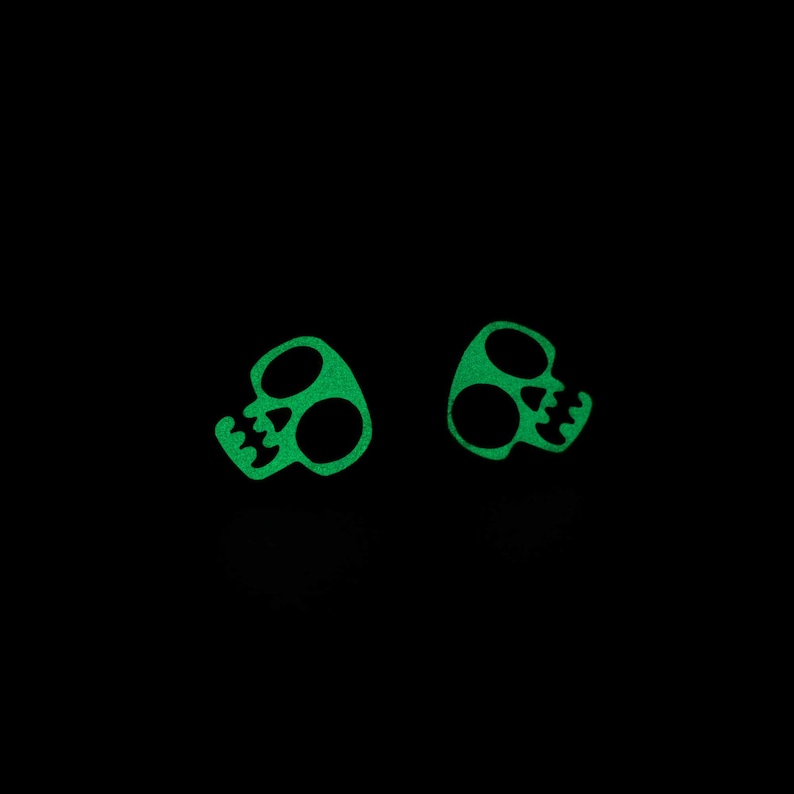 May include: Two pairs of neon green skull-shaped earrings against a black background. The earrings have large eye sockets and a jagged jawline. The earrings are a fun and edgy accessory.