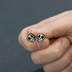 May include: A pair of black and white skull stud earrings. Each earring features a small, cartoon-style skull design with white details on a black background. The earrings are held in a hand against a gray background.
