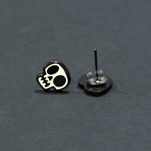 May include: Black and white skull stud earrings. The earrings feature a black skull design with white accents. The post is silver-colored. The earrings are set against a dark gray background.