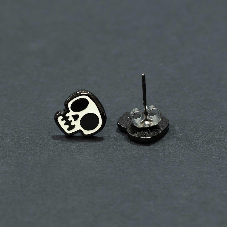 May include: Black and white skull stud earrings. The earrings feature a black skull design with white accents. The post is silver-colored. The earrings are set against a dark gray background.