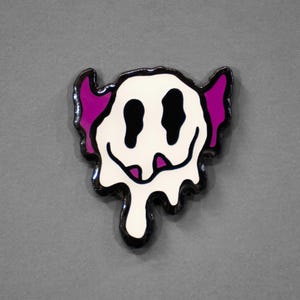 May include: A white, black, and purple enamel pin with a melting smiley face design. The pin features black eyes and a smiling mouth, with purple accents on the ears. The edges are outlined in black.