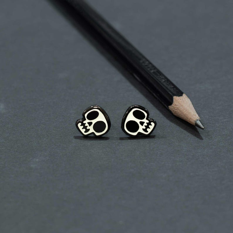 May include: A pair of black and white skull-shaped stud earrings. The earrings have a glossy black outline with a white skull design. A black pencil is in the background, resting on a dark gray surface.