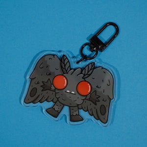 May include: A clear acrylic keychain featuring a cartoon moth with gray wings, body, and antennae. The moth has large red eyes and a black clasp. The keychain is set against a blue background.