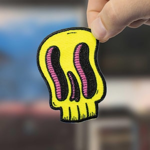 May include: A yellow skull patch with black eye sockets and a pink interior. The patch has a black outline and is held by a hand. The skull design is cartoonish and has a unique, wavy shape. The patch is likely for clothing or accessories.