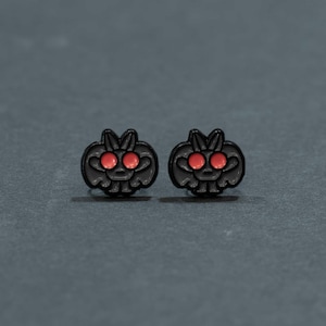 Puede incluir: Un par de pendientes de botón con forma de pequeñas criaturas estilizadas con cuerpos negros y ojos redondos, prominentes y de color rojo brillante. El diseño presenta una silueta simétrica y redondeada con pequeñas orejas, hecho en metal oscuro.