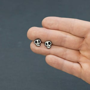 May include: Two small, black skull earrings with white details are displayed. The earrings are held in a person's hand against a dark gray background. The skull design features eye sockets, a nose cavity, and a jawline.