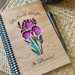 May include: A wooden journal with a spiral binding. The cover is engraved with the text "The Earth Laughs in Flowers!" and a cutout design of a purple iris.