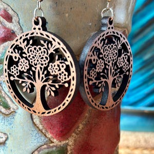 May include: A pair of wooden earrings featuring a tree of life design. The earrings are circular with a dark brown outer ring and a lighter brown tree design cut out of the center. The earrings hang from silver-colored hooks.