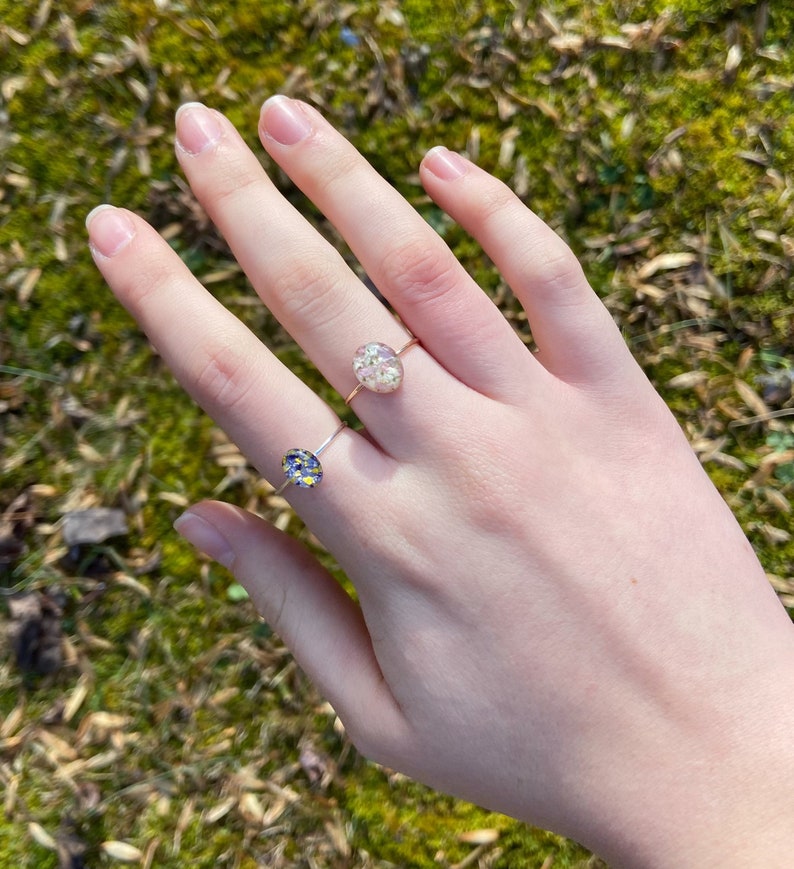 Pu&ograve; includere: Due anelli d'oro con bande delicate e montature in resina ovali. L'anello pi&ugrave; grande presenta un motivo floreale bianco, mentre l'anello pi&ugrave; piccolo ha un motivo floreale blu e giallo.