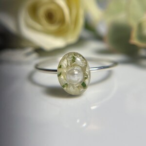 Pu&ograve; includere: Un anello d'argento con una perla bianca e fiori pressati verdi e bianchi incapsulati in resina trasparente.