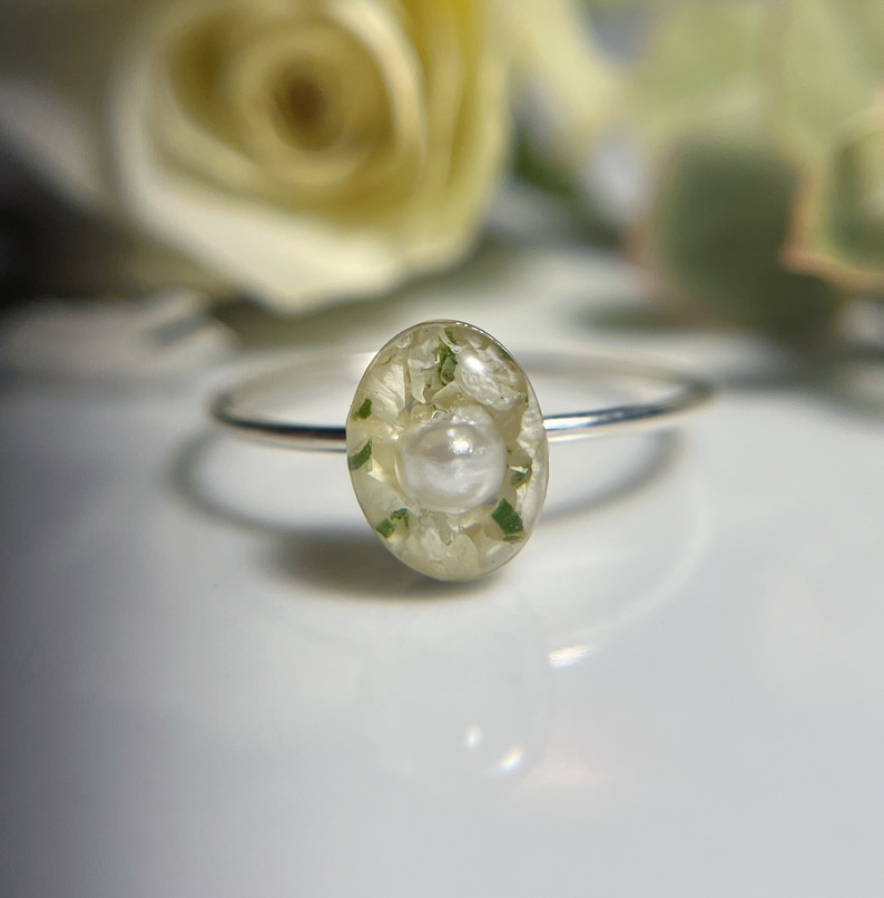 Pu&ograve; includere: Un anello d'argento con una perla bianca e fiori pressati verdi e bianchi incapsulati in resina trasparente.