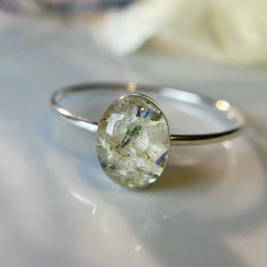 Pu&ograve; includere: Un anello d'argento con una montatura ovale in resina bianca contenente fiori pressati e brillantini dorati.