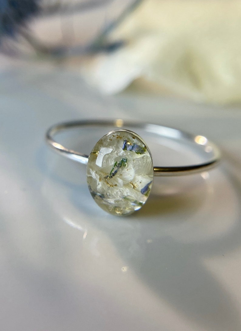 Pu&ograve; includere: Un anello d'argento con una montatura ovale in resina bianca contenente fiori pressati e brillantini dorati.