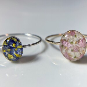 Pu&ograve; includere: Due anelli d'argento con inserti ovali in resina. L'anello a sinistra presenta un motivo floreale blu e giallo. L'anello a destra presenta un motivo floreale rosa e bianco.