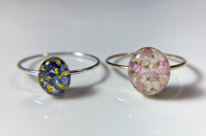Pu&ograve; includere: Due anelli d'argento con inserti ovali in resina. L'anello a sinistra presenta un motivo floreale blu e giallo. L'anello a destra presenta un motivo floreale rosa e bianco.