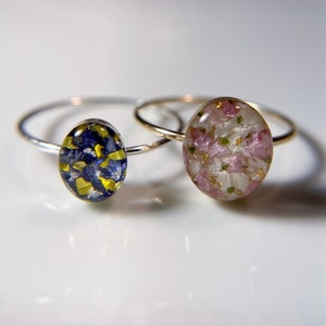 Pu&ograve; includere: Due anelli d'argento con montature in resina ovali. Un anello presenta un design floreale blu e giallo, mentre l'altro anello presenta un design floreale rosa e bianco.