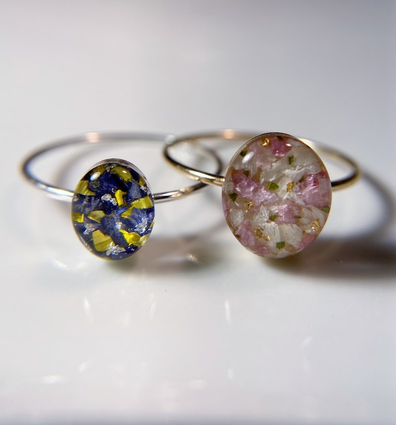 Pu&ograve; includere: Due anelli d'argento con montature in resina ovali. Un anello presenta un design floreale blu e giallo, mentre l'altro anello presenta un design floreale rosa e bianco.