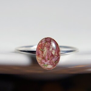 Pu&ograve; includere: Un anello d'argento con una pietra ovale in resina rosa e bianca. La pietra &egrave; piena di piccoli pezzi di fiori secchi.