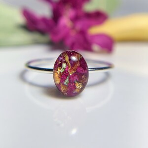 Pu&ograve; includere: Un anello d'argento con una montatura rotonda in resina trasparente che contiene fiori rosa e dorati pressati.