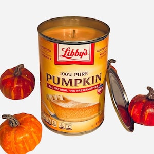 May include: A candle in a metal can with a pumpkin pie design. The can is labeled "Libby's 100% Pure Pumpkin All Natural - No Preservatives". There are three small orange pumpkins on a white background.