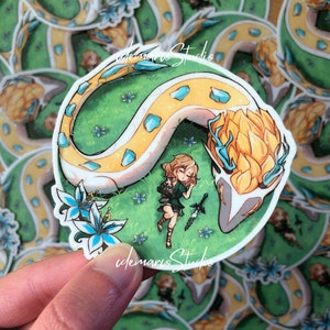 Op de afbeelding: Een sticker met een cartoonillustratie van een witte en gele draak die zich om een ​​slapend meisje in een groen veld krult. De draak heeft blauwe accenten en het meisje houdt een zwaard vast. De sticker heeft een witte rand en de tekst "idemarro Studio" staat onderaan gedrukt.